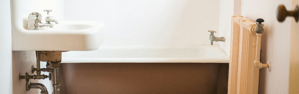 Ein kleines Badezimmer mit einer freistehenden Badewanne, einem Waschbecken mit alten Wasserhähnen und einem Heizkörper an der Wand. Die Sanitäranlagen wirken altmodisch und der Raum ist mit weißen Wänden ausgestattet.