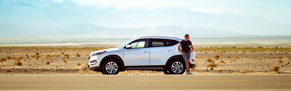 Ein Mann steht neben einem silbernen SUV am Straßenrand in einer trockenen, wüstenartigen Landschaft. Im Hintergrund sind Berge und ein blauer Himmel zu sehen.