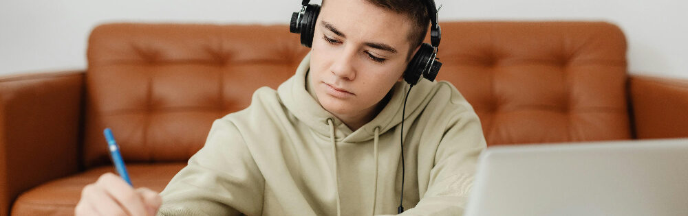 Ein junger Mann sitzt auf einem Sofa und trägt Kopfhörer. Er konzentriert sich auf das Schreiben mit einem Stift auf Papier, während vor ihm ein Laptop steht.